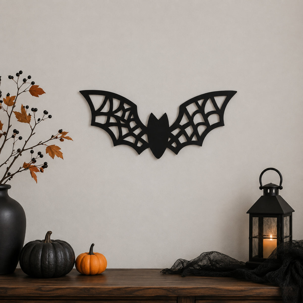Black bat wall decor with intricate web-like cutout details, displayed on a beige wall above a wooden mantel decorated with pumpkins, a black vase with autumn branches, and a black lantern.