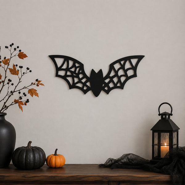 Black bat wall decor with intricate web-like cutout details, displayed on a beige wall above a wooden mantel decorated with pumpkins, a black vase with autumn branches, and a black lantern.