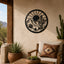 A circular black metal wall art piece featuring a desert landscape with a saguaro cactus, mountains, and a rising sun, mounted on a beige stucco wall in a patio setting with a wooden chair and potted plants.