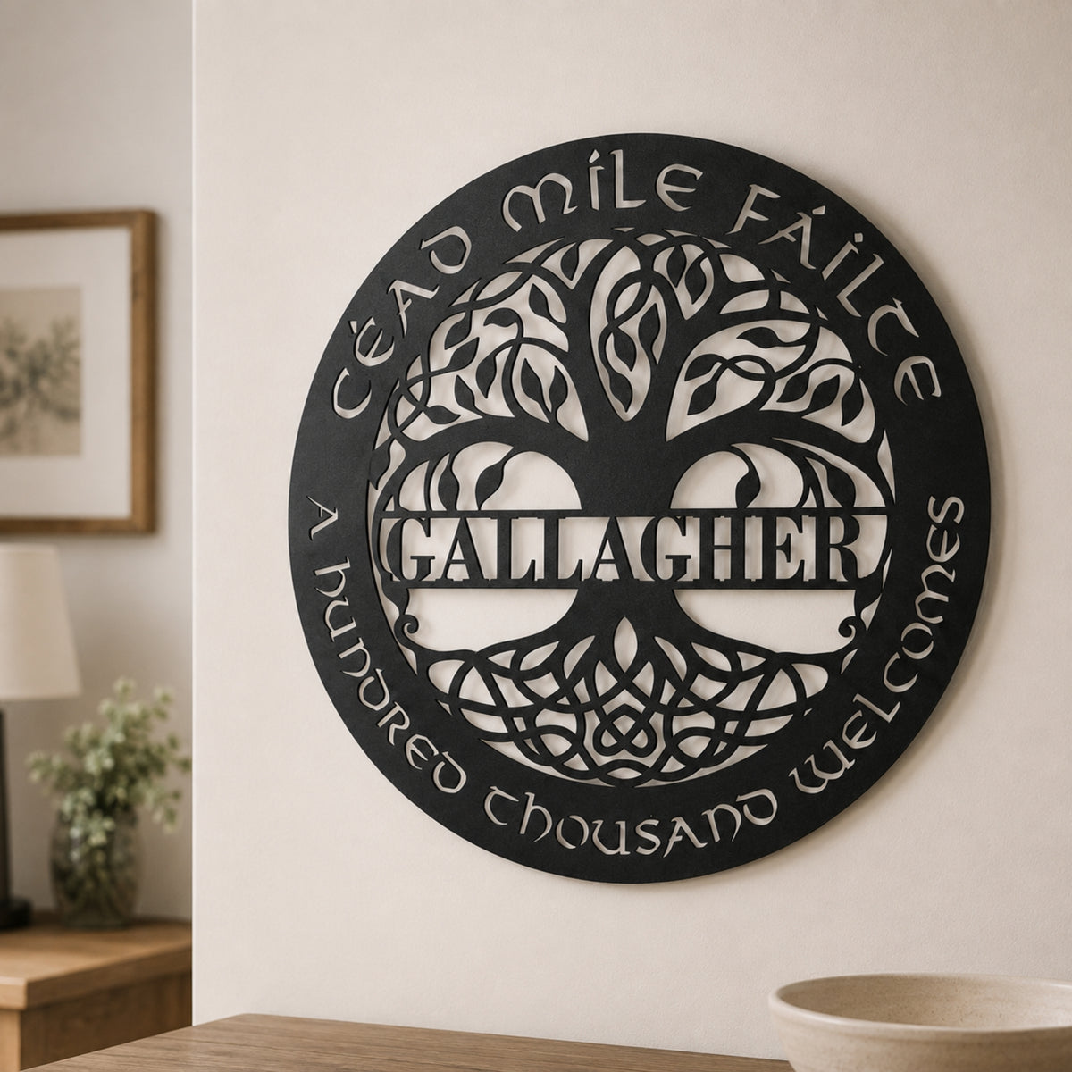 A circular black metal wall sign mounted on a beige wall, featuring a tree of life design with the name 'GALLAGHER' across the center and the phrases 'CÉAD MILE FÁILTE' and 'A HUNDRED THOUSAND welcomes' around the edge. The sign is shown in a styled interior setting with a lamp, framed picture, and table visible.