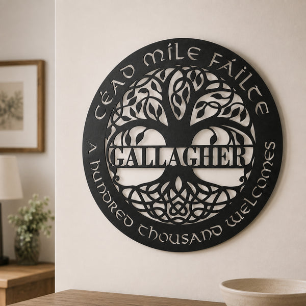 A circular black metal wall sign mounted on a beige wall, featuring a tree of life design with the name 'GALLAGHER' across the center and the phrases 'CÉAD MILE FÁILTE' and 'A HUNDRED THOUSAND welcomes' around the edge. The sign is shown in a styled interior setting with a lamp, framed picture, and table visible.
