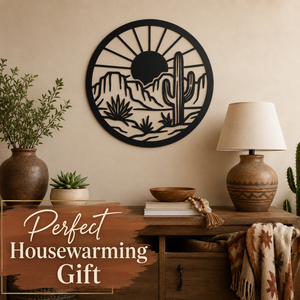 A round black metal wall art piece featuring a desert landscape with a saguaro cactus, mountains, and a sun, displayed on a beige wall above a wooden console table with a lamp, plants, and a throw blanket. A text overlay in the bottom left reads 'Perfect Housewarming Gift'.