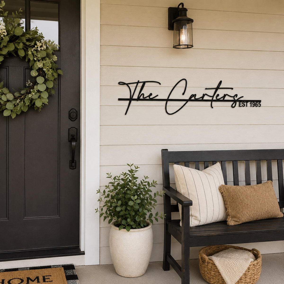 Black metal script wall art reading 'The Carters Est 1955' mounted on a beige wood-paneled wall on a front porch, next to a dark bench with pillows, a potted plant, and a dark door with a wreath.
