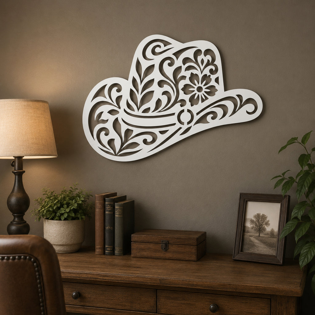 A white decorative wall sign shaped like a cowboy hat with intricate floral cutout patterns, mounted on a brown wall above a wooden console table in a styled room setting.