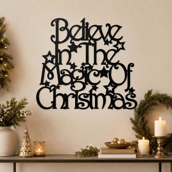Black metal wall art with the phrase 'Believe in the Magic of Christmas' in decorative script and star motifs, displayed on a beige wall above a styled holiday console with candles, greenery, and a small tree.